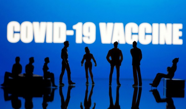 Small toy figures are seen in front of a Covid-19 Vaccine logo in this illustration taken