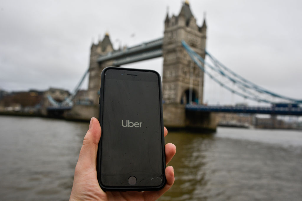 The Uber Logo is Displayed on a Phone in Front of Tower Bridge in London