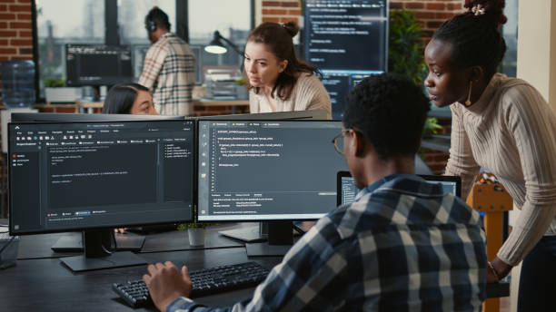Mixed team of artificial intelligence cloud programers talking about programming in front of multiple screens stock photo
